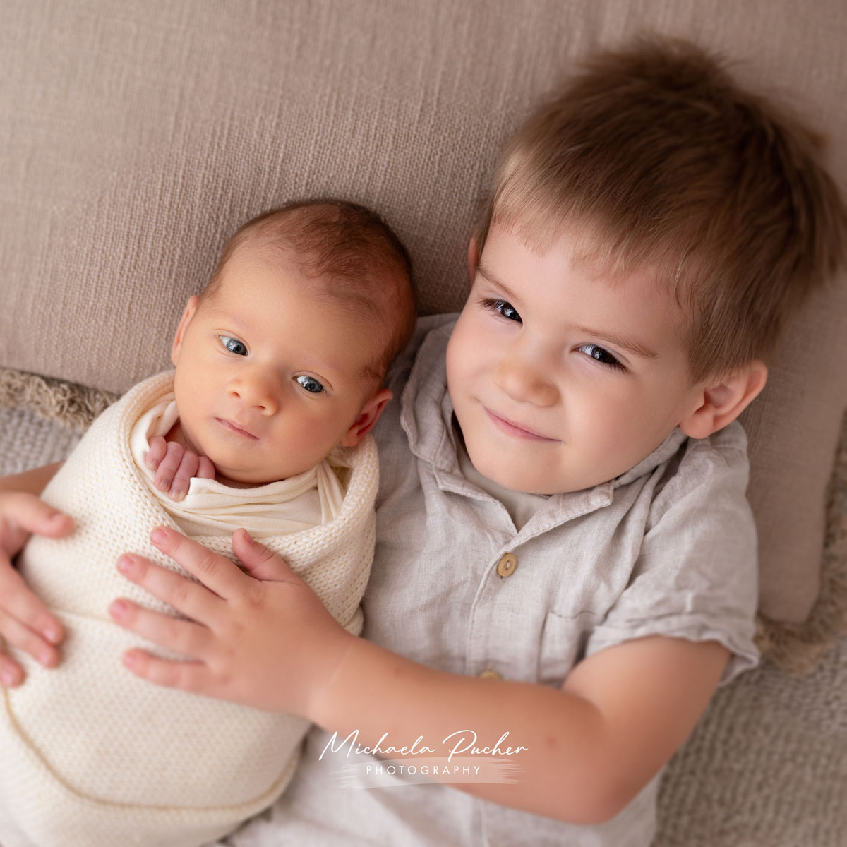 Kleinkind hält sein eingewickeltes Neugeborenes im Arm, beide liegen auf einer weichen Unterlage in warmen Naturtönen.